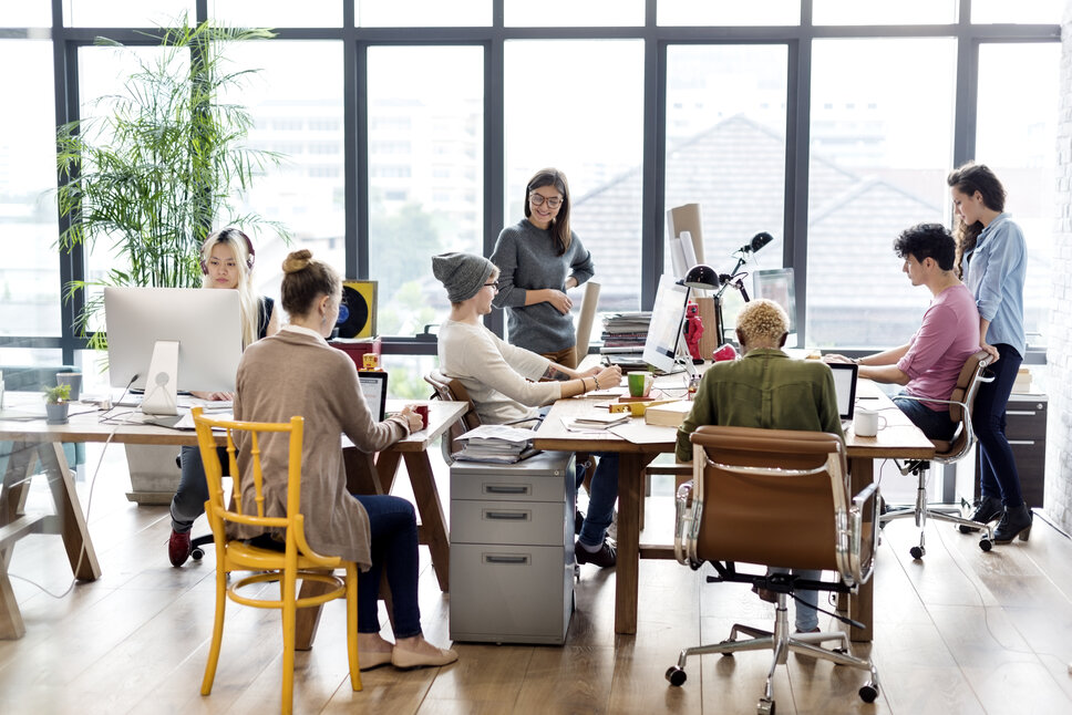 In einem modernen, offenen Büro sitzen viele Mitarbeiter:innen an einem Tisch. 