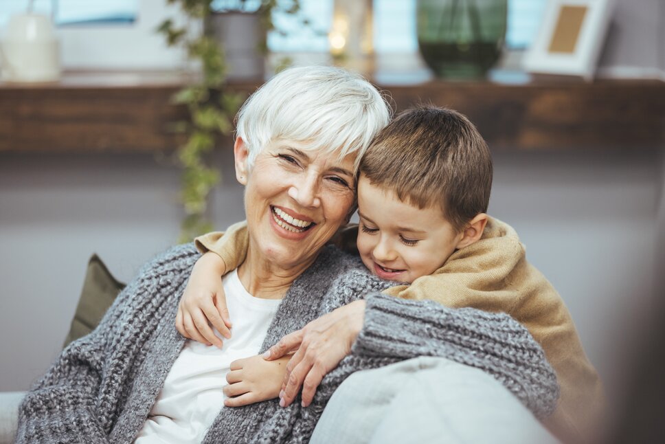 Eine ältere Dame mit weißen Haaren sitzt gemeinsam mit ihrem jungen Enkelkind auf dem Sofa. 