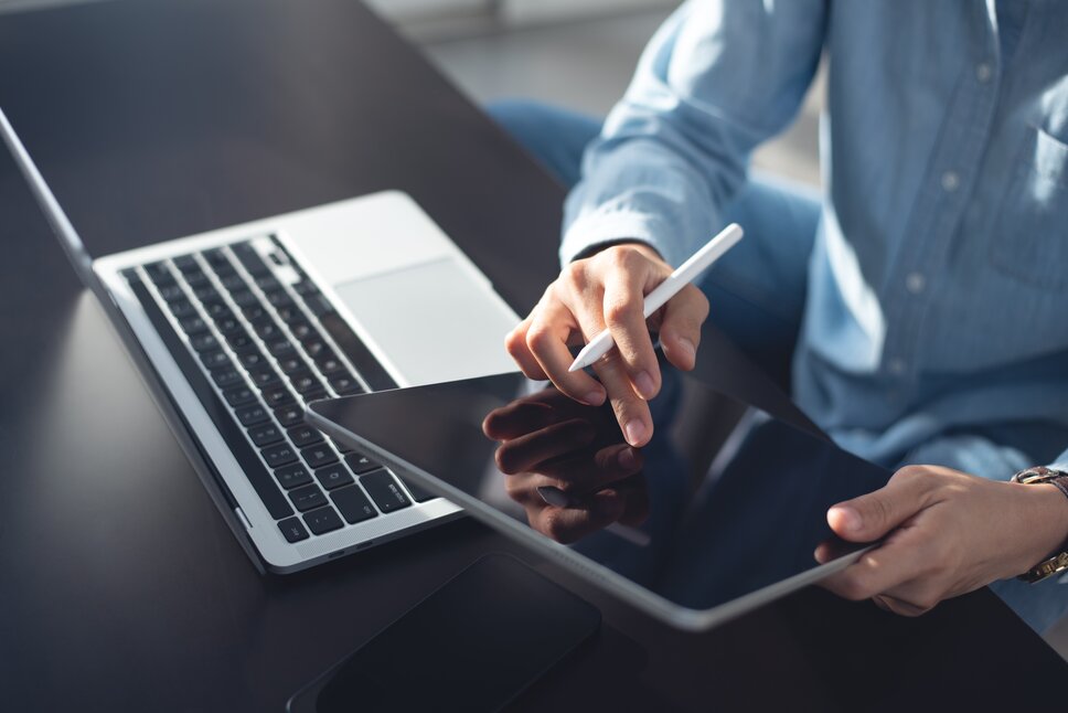 Eine Nahaufnahme von einem Mann im Business-Outfit, der vor einem Laptop sitzt und ein Tablet in den Händen hält.